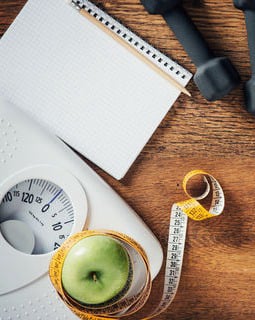 An overhead view of a scale, green apple, measuring tape, and fitness equipment on a wooden table