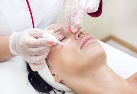 Skincare professional wearing clear gloves applies a white cream or treatment mask to a client's under-eye area using a small applicator tool during a customized facial treatment
