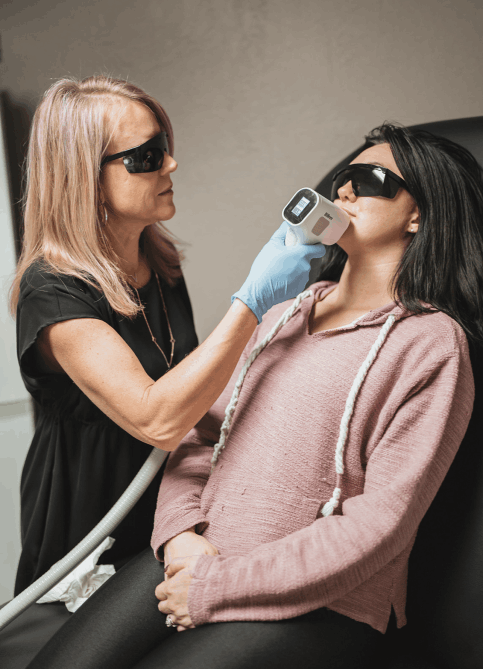 A professional and client wearing safety goggles while a handheld laser device is used for a hair removal treatment on the chin
